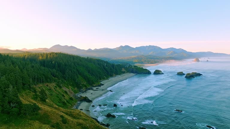 Aerial at golden hour sunrise tracking shot of Crescent Beach, Bird Rocks with Haystack Rock and Cannon Beach on the Oregon coast on a beautiful blue-sky summer day, mountains in the distance.