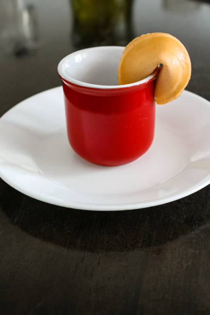 A little red mug has a fortune cookie attached to the edge.  It sits on a white plate on a black table