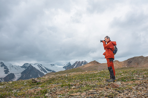 Man in red with camera shoots high snowy mountain range with sharp tops under gray cloudy sky. Tourist photographs dramatic landscape with large snow mountains with pointed peaks at changeable weather
