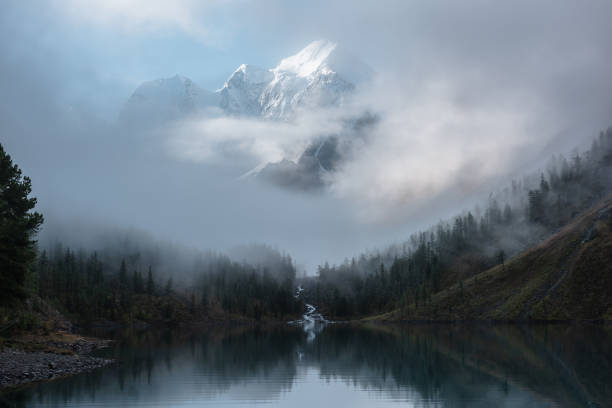 istock Tranquil scenery with snow castle in clouds. Mountain creek flows from forest hills into glacial lake. Snowy mountains in fog clearance. Small river and coniferous trees reflected in calm alpine lake. 1480878573