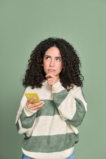 Young thoughtful unsure latin woman holding hand on chin using mobile phone isolated on green background. Female model with cell thinking, choosing, feeling doubt or making decision concept. Vertical.