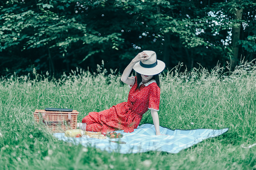 A beautiful young caucasian girl in a red dress covering her face with a straw hat sits on a blue bedspread with a wicker square basket and fruits laid out in plates in a clearing in a public park, side view close-up.Outdoor picnic concept.