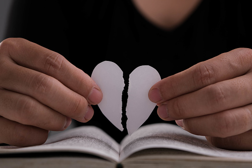 Female hand holding a half-torn white paper heart over an open book on the table. White paper heart torn in half in hand