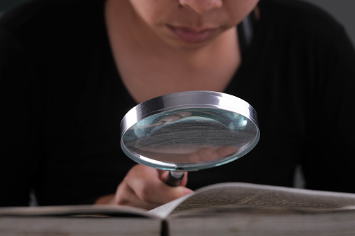 Close-up of a woman looking through a magnifying glass at a textbook. Magnifying glass in hand and open book on table. Education and research concept.