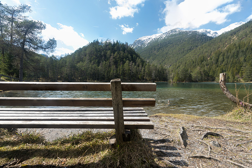 Landscape wooden bench at the idyllic mountain lake Fernsteinsee in Tyrol Austria to relax with partly still snow covered mountains hills and bluish green clear water clouds sky break and relaxation