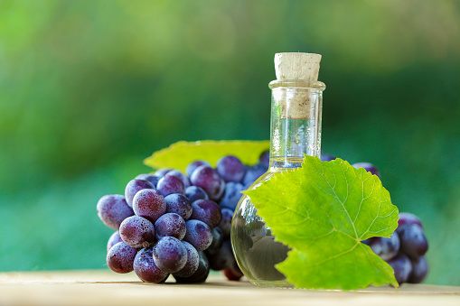 Grape vinegar in glass round bottle, grapes bunches with green leaves on a wooden saw cut on a blurred green garden background.Organic Natural Bio Grape Seed Oil.