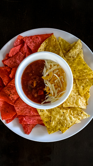 White plate of red and yellow tortilla chips with a bowl of cheese and salsa in the middle.  On black background
