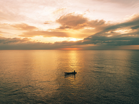 Fisherman sitting a small thai fishing longtail boat fishing during sunset in the Andaman Sea close to Khao Lac Island under moody orange dusk skyscape. Drone Point of View. Moody sunset over the Andaman Sea. Khao Lak Island, Andaman Sea, Phuket, Thailand, Southeast Asia.