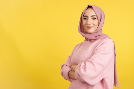 Muslim woman standing with arms crossed looking at camera in studio with yellow background