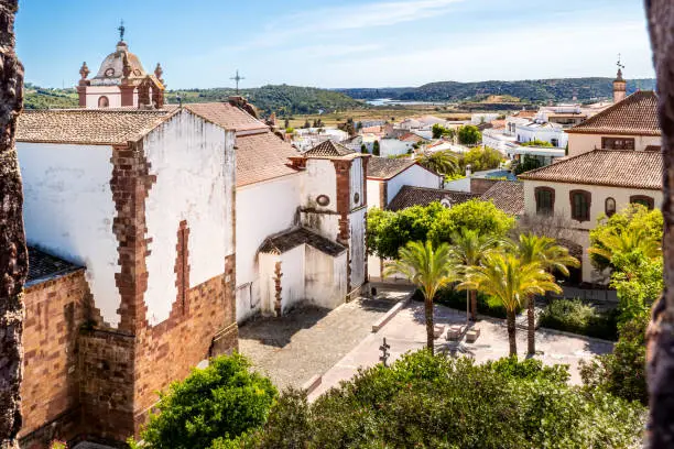 Photo of From castle of Silves, the Silves cathedral and its lateral square can be seen in front of Cidade de Silves rooftops, with palm trees and lush surroundings leading to Arade river under a sunny sky.