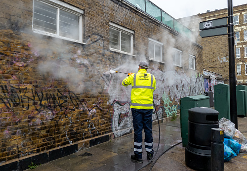 London. UK- 04.08.2022. A Tower Hamlet Council worker cleaning graffiti off a building with a jet power wash.
