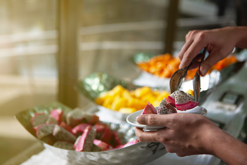 Restaurant guest takes dragon fruit for breakfast or dessert at a luxury hotel buffet. He takes the fruit from the large bowl with tongs and pours it into his saucer