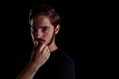 Close up of young adult male looking sinister or contemplative. Color dark tones for dramatic effect, dark and moody series. Concept image for corporate scheming. Thumb rubbing lips.