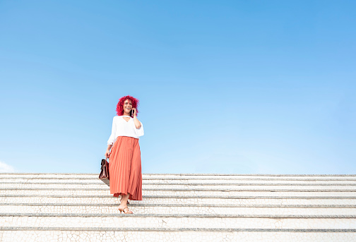 horizontal portrait of a happy fashionable businesswoman with red afro hair holding handbag and walking down white stairs while talking on smartphone against blue sky