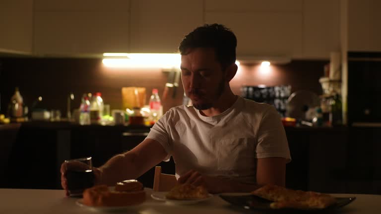 Front view of handsome young man feeling hungry, eating hamburger and drinking unhealthy beverage sitting alone at table in evening or night