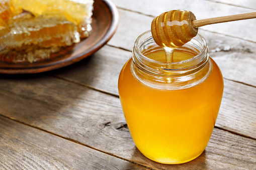 honey pouring from dipper in glass jar and honeycomb on clay plate on wooden table