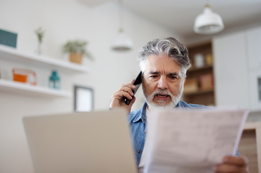 Stressed mature man holding paper and talking on phone at home. Discussing document
