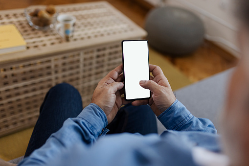 Male tech user relaxing on sofa holding smartphone mockup blank white screen. Man customer using cell phone mobile social media or shopping app, reading news, sport betting. Over shoulder view