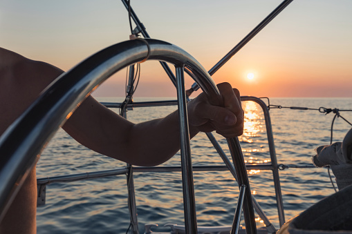 Winde of a sailboat with attached crank in the evening, rope holding the sail mast, empty boat deck, sea with island and beautiful sunset sky in the background