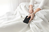 Mockup image of a woman in bed in her bedroom holding black smartphone mobile phone. The girl freelancer  in a white T-shirt is working of a bed with white bed sheets.Employee works during sick leave.