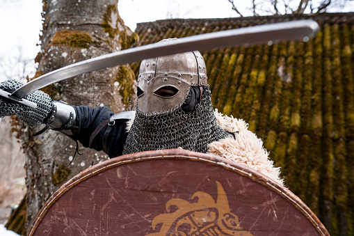 Back view of warrior man wearing iron helmet and red cloak holding sword, soldier looking and turned back. Male wearing like spartan or antique roman at field on sunset.