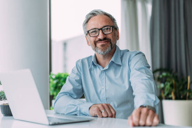 úspěšný podnikatel v moderní kanceláři pracující na notebooku. - pohled do objektivu - stock snímky, obrázky a fotky