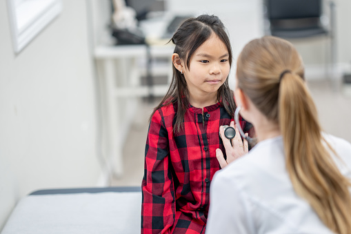 A female doctor leans in closely as she listens to the heart of a young patient.  The doctor is dressed professionally in scrubs and the little girl is seated on the  exam table as she sits still for the exam.