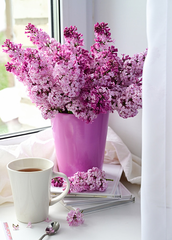 The postcard is beautiful. A mug of tea, an old book and a bouquet of purple lilac. A beautiful still life. Spring time. The concept of Good morning.
