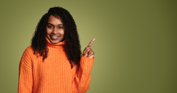 Happy African American female in orange sweater smiling and pointing up to copy space while standing against green background