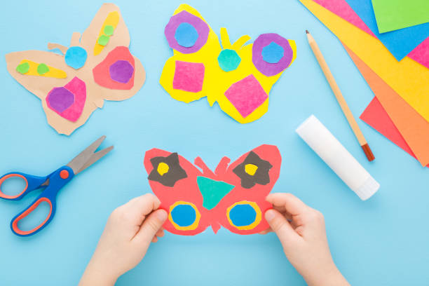 istock Little child hands holding colorful butterfly shape from paper on light blue table background. Pastel color. Making decoration elements. Closeup. Point of view shot. Top down view. 1480984942