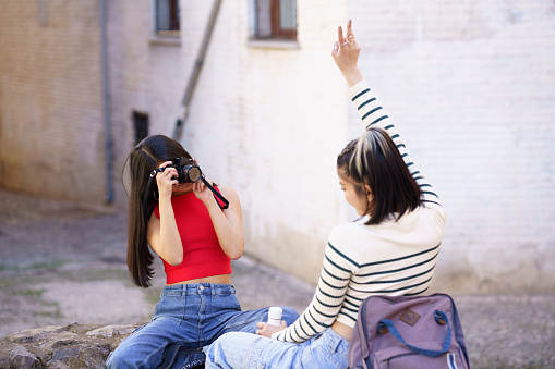 Young female photographer taking photo of girlfriend showing peace gesture on city street during vacation