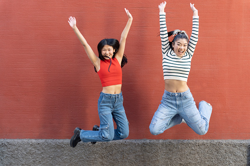 Full body of happy ethnic female friends wearing jeans enthusiastically jumping up with raised arms against red wall looking at camera