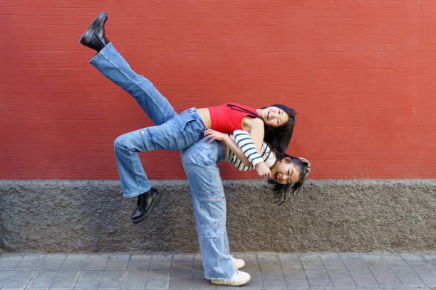 Side view of cheerful Asian woman in casual clothes lifting smiling friend on back while standing against red wall