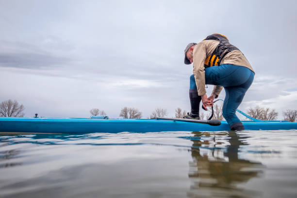 senior male is attaching a safety leash before paddling a stand up paddleboard senior male is attaching a safety leash before paddling a stand up paddleboard on a lake in early spring, frog perspective from an action camera at water level paddleboard surfing water sport low angle view stock pictures, royalty-free photos & images