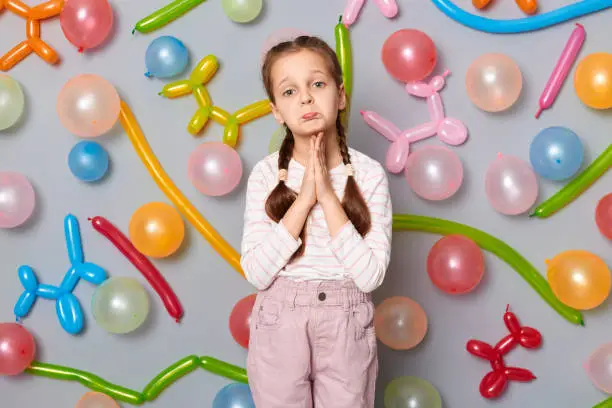 Pleading child. Sad upset little girl with pigtails standing against gray wall decorated with colorful balloons keeps hands in praying gesture asking to forgive.