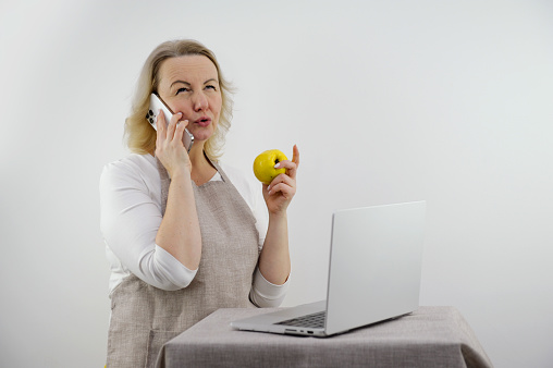 woman talking on the phone is upset and displeased in her hands she has apple dietetics proper nutrition menu recipes in front of laptop in hands phone apron adult middle-aged woman blonde