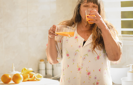 Caucasian mature senior woman drinks a glass of freshly squeezed natural orange juice with pulp.breakfast concept and wake up in the morning.very bright and luminous home kitchen.