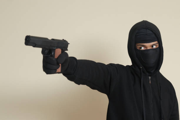 Mysterious man wearing black hoodie and mask holding a pistol, shooting with a gun. Isolated image on gray background stock photo