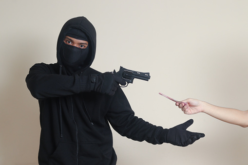 Portrait of mysterious man wearing black hoodie and mask doing shooting with gun while extorts rupiah money banknote from victim. Isolated image on gray background