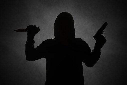 Mysterious man wearing black hoodie holding a pistol, shooting with a gun while holding a knife. Silhouette and dark concept image