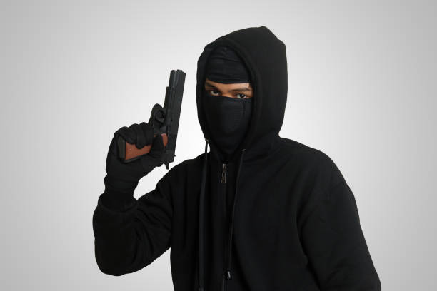 Mysterious man wearing black hoodie and mask holding a pistol, shooting with a gun. Isolated image on gray background stock photo