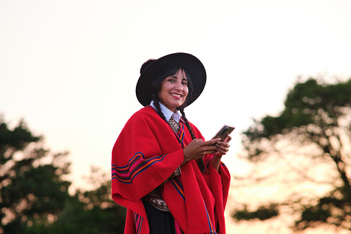 Native woman in traditional clothes smiling at the camera while using a mobile in the mountain during sunset