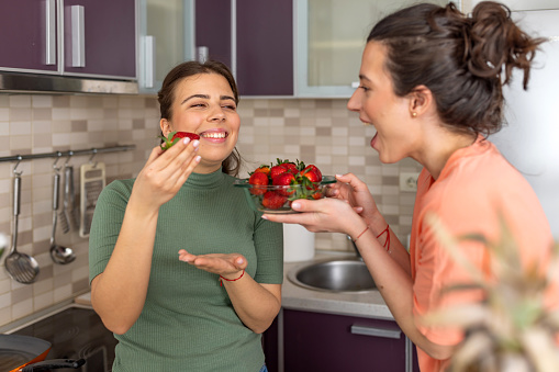 Two friends are standing in the kitchen and enjoying fresh strawberries. They are both smiling and chatting while taking turns eating the berries