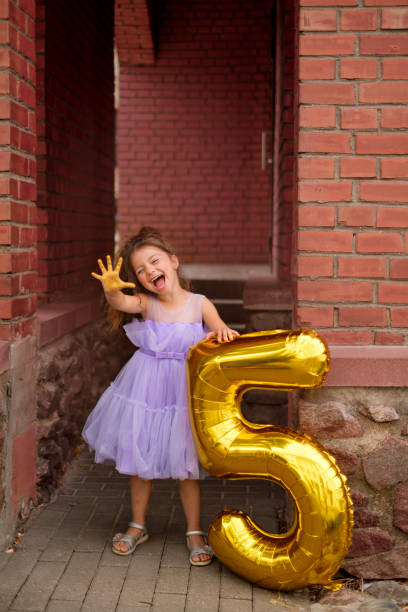 紫色のドレスを着た女の子が、手に5本の指を見せて陽気に笑っています。ゴールデンナンバーファイブの形をした風船が子供の隣に立っています。誕生日パーティー。 - merrily ストックフォトと画像