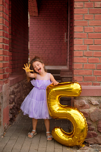 A girl in a purple dress laughing merrily showing five fingers on her hand. A balloon in the shape of the Golden number Five stands next to the child. Birthday party.