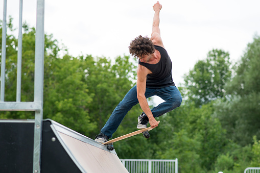 Athletic man with modern curly hair and wearing a sleeveless black shirt and blue jeans is doing a stall trick at the top of a half pipe in a skateboard park outdoors.