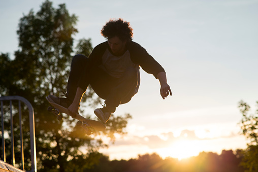 Silhouette of a talented, athletic young man with curly hair jumping in the air on his skateboard off of the top of a half pipe skateboard ramp at sunset with the sky and sun behind him.