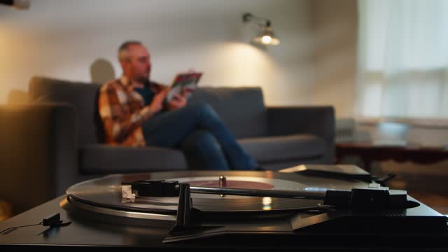Man on couch reads a book and listens to a retro turntable with a vinyl record.