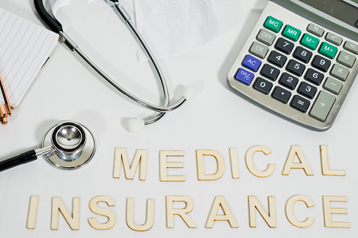 The words medical insurance are spelled out in wooden letters on a white background. These words are surrounded by a stethoscope, calculator, and papers.