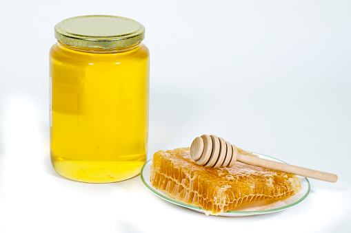 A jar of honey sits on a white background. Next to it, a plate of honeycomb with a honey dipper on top of it.